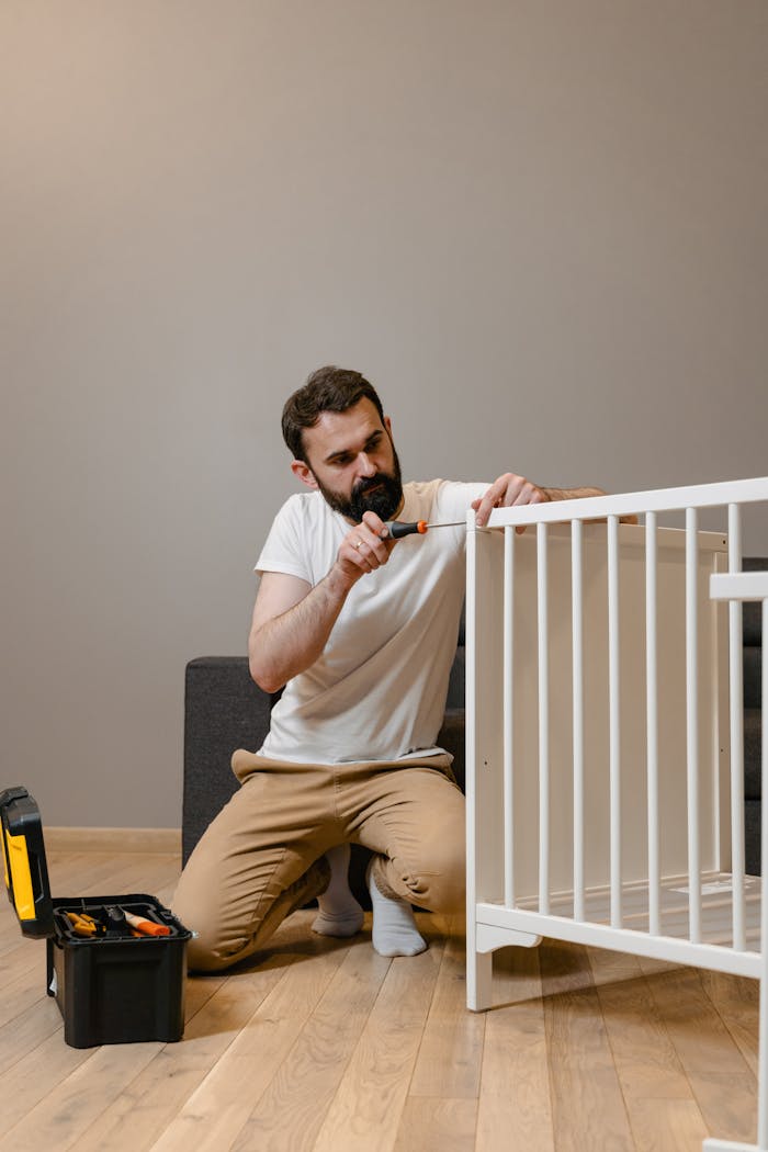 Bearded man assembling a white crib with tools on the wooden floor indoors.